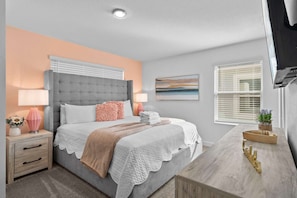 A bedroom with a grey tufted headboard, a white bedspread, a pink throw, and a bedside table with a lamp and a vase of flowers. - 5BR Townhome w/ Pool at Champions Gate (Davenport)