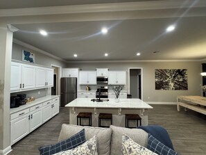 A modern kitchen with white cabinets, a central island, and a dining area with a bench. - Game Day Getaway / Near Sports Complex / Sleeps 10 / 4BR/3BA (Youngsville)