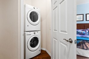 A laundry room with stacked washing machines and a door leading to a bedroom with a bed and a wall-mounted painting. - 4BR Resort Townhome 15 Min From Disney (Kissimmee)