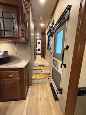 A narrow hallway with wooden cabinets, a kitchen area with a countertop and appliances, and a staircase leading to a laundry room.