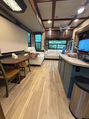 A compact RV interior with a dining area, seating, a kitchenette, and a television. - Experience Luxury RV Living - Smoky MTN & River Getaway in Spacious 5th Wheel (Walland)