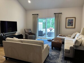 Ein modernes Wohnzimmer mit einer Couch, Sesseln, einem Couchtisch, einem Fernseher und einer Schiebetür zu einem Außenbereich.