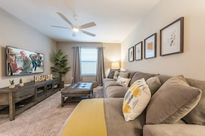 A living room with a sofa, a coffee table, a TV, a ceiling fan, and framed pictures on the wall. - Luxury 6-bedroom Retreat w/ Heatable Pool (Kissimmee)