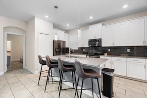 A modern kitchen with white cabinets, a central island with bar stools, a stainless steel refrigerator, and a black microwave. - Storey Lake 5BR Themed Villa 10 min to Disney (Kissimmee)