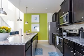 A modern kitchen with dark cabinets, a granite countertop, a stainless steel refrigerator, and a microwave. There are two pendant lights, a sink, and a large window with a view of the outside. - 6BR Disney Retreat w/ Pool & Themed Rooms (Davenport)