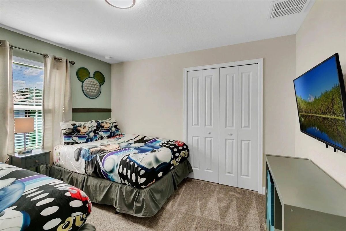 Un dormitorio con una cama con colcha temática de Mickey Mouse, una ventana con persianas, una puerta y un televisor sobre un mueble.