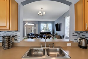 A modern kitchen with a double sink, a dining area with a black leather couch, and a large window with sheer curtains. - Family 6BR Villa w/ Marvel Game Room & Pool (Kissimmee)