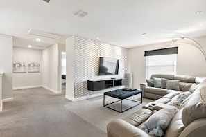 A modern living room with a sectional sofa, a coffee table, a TV mounted on a geometric-patterned wall, and artwork on the walls. - 6BR Storey Lake Home, Private Pool (Kissimmee)