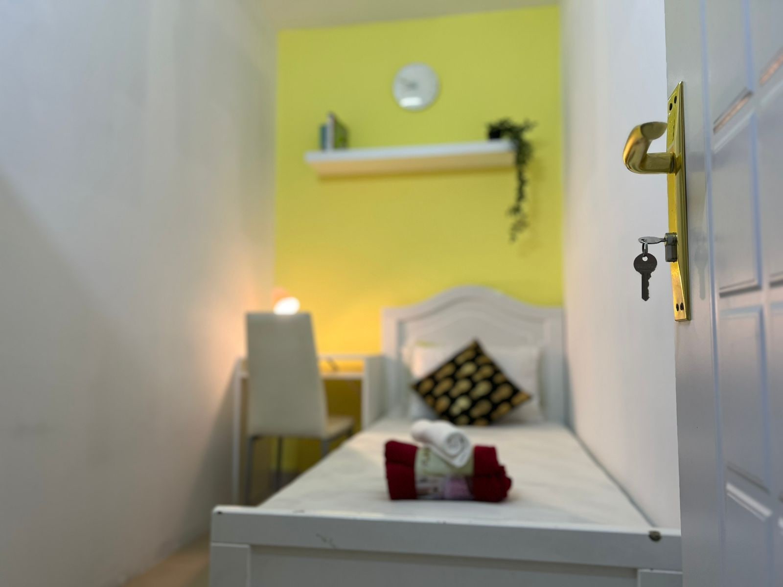 Een kleine kamer met een bed, nachtkastje, een klok aan de muur en een plankje met een plant.