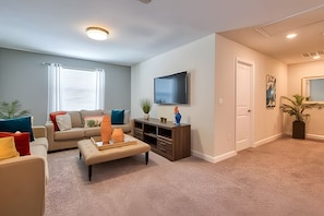 A living room with a sofa, ottoman, TV, and entertainment center. - 6BR Pool Home w/ Mickey + Avengers Storey Lake (Kissimmee)