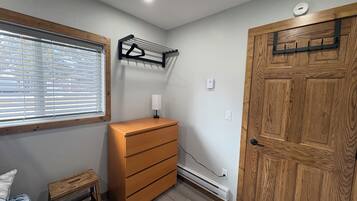 Un dormitorio con cómoda de madera, mesita de noche, perchero, ventana con persianas y puerta.