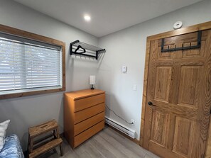 A bedroom with a wooden dresser, a bedside table, a hanging rack, a window with blinds, and a door.