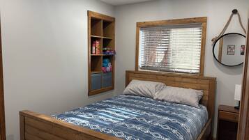 Un dormitorio con cama de madera, estante empotrado con espacio de almacenamiento, ventana con persianas y un espejo redondo en la pared.