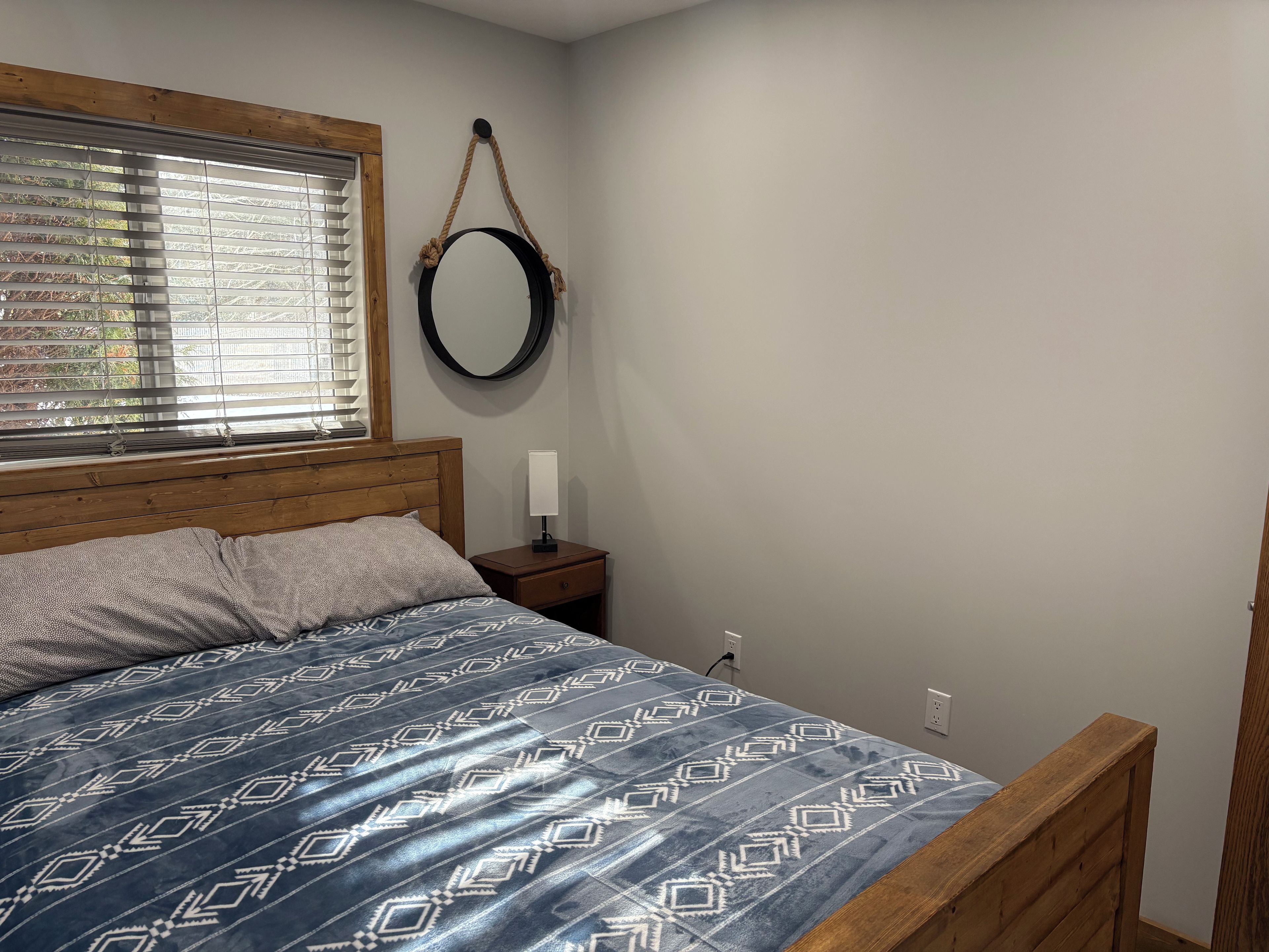Une chambre à coucher avec une tête de lit en bois, une couvre-lit à motifs, une table de chevet avec une lampe et une fenêtre avec des stores.