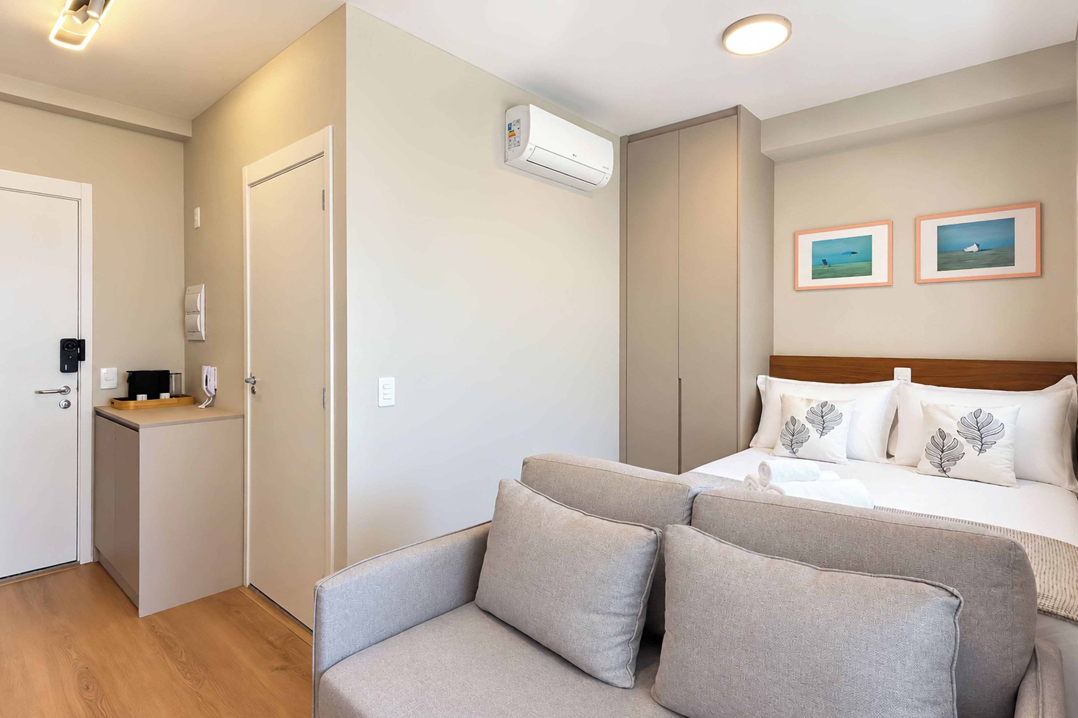 A hotel room with a bed, a sofa, an air conditioning unit, a small kitchenette, and two framed pictures on the wall.