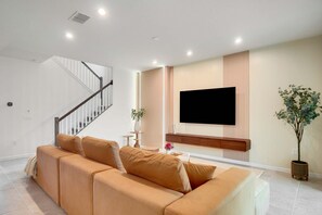 A modern living room with a beige sofa, a flat-screen TV mounted on the wall, a staircase, and a potted plant. - 7BR w/ Private Pool & Game Room (Kissimmee)