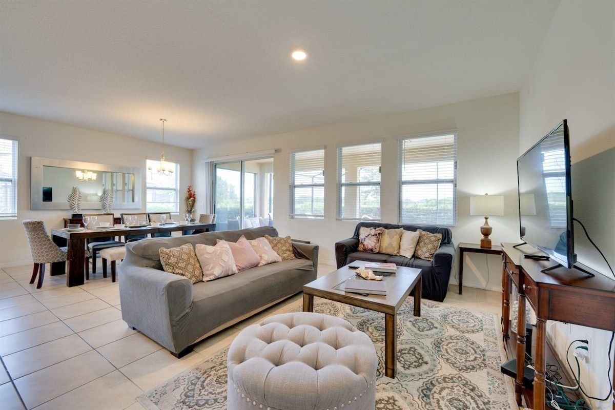 A spacious living room with a grey sofa, a coffee table, and a TV. There is a dining area with a table and chairs, and a kitchen in the background.