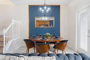 A dining area with a round table set for four, surrounded by wooden chairs. A chandelier hangs above the table, and a mirror is mounted on the wall. - Modern 5BR Villa w/ Private Pool at Windsor (Kissimmee)