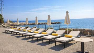 A beachside lounge area with sun loungers and umbrellas, a round table, and a view of the sea.