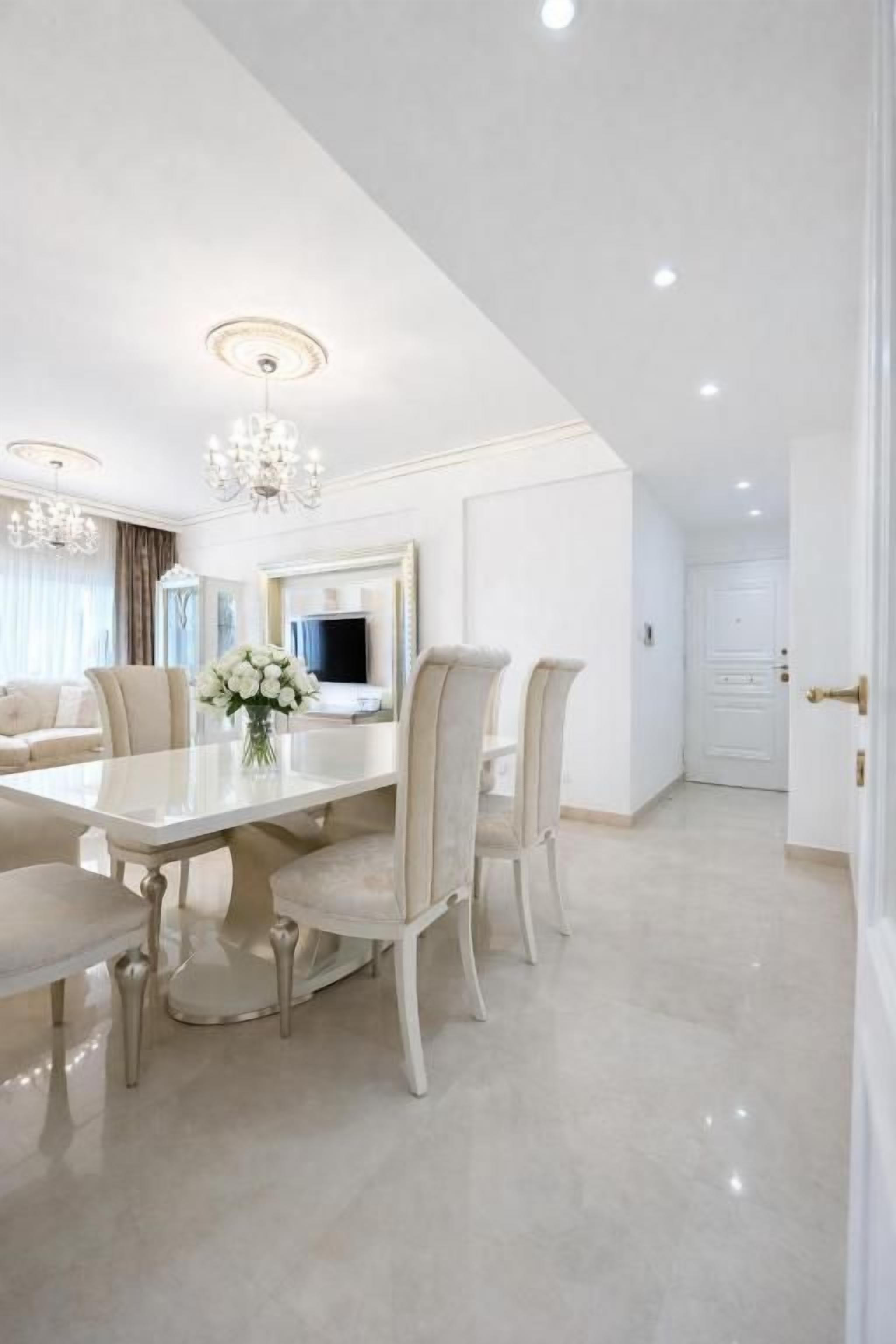 Un coin repas avec une table et des chaises blanches, un lustre et un miroir au mur.