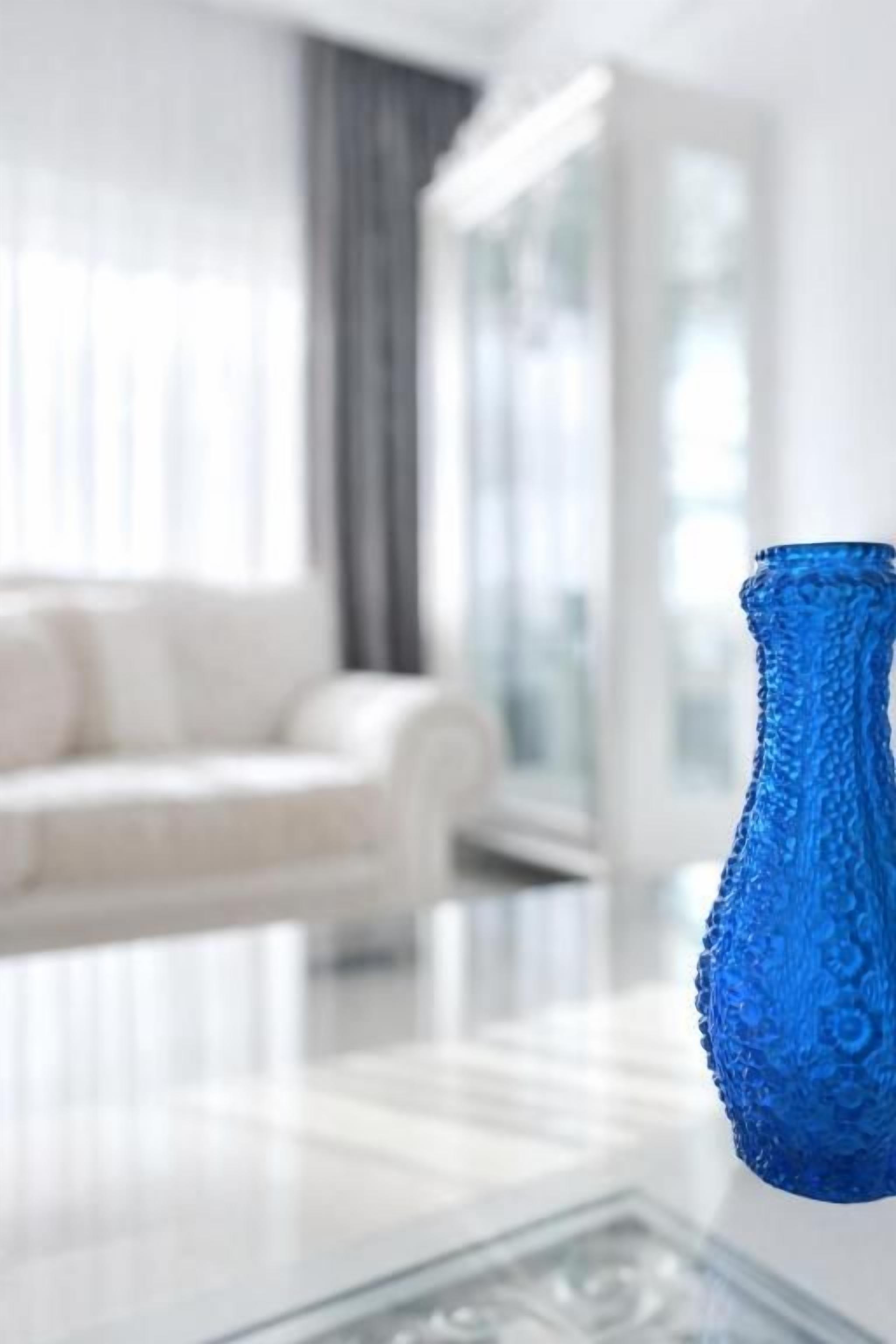 A blue textured vase on a glass table in a modern living room with a white sofa and a glass door in the background.