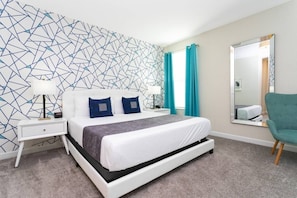 A modern hotel room with a geometric-patterned wall, a large bed with white bedding and two blue pillows, a side table with a lamp, a teal chair, and a large mirror on the wall. - 6BR Villa w/ Pool, Spa & Resort Close to Disney (Kissimmee)