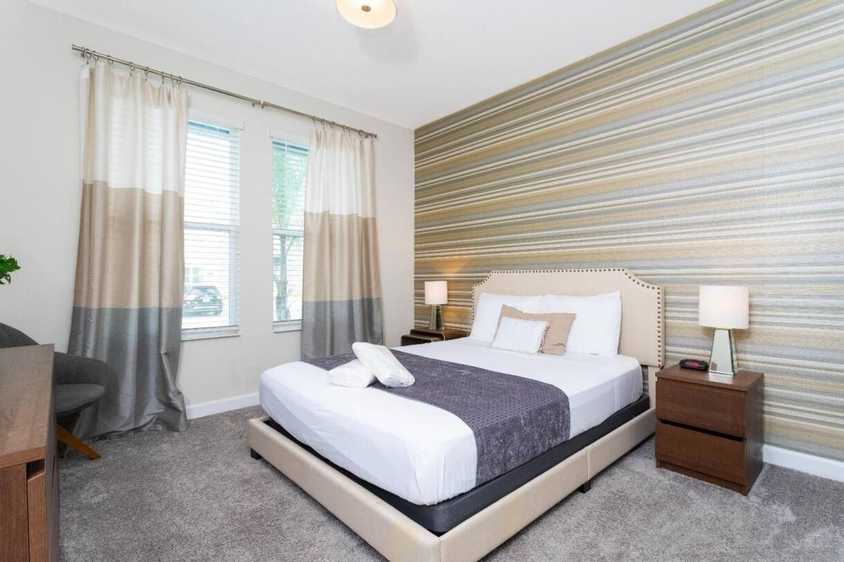 A hotel room with a large bed, two bedside tables with lamps, a grey sofa, and a striped wall.