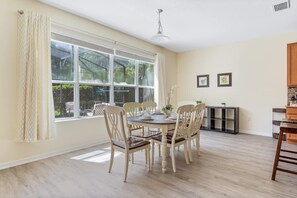 Un comedor con una mesa redonda y seis sillas, una ventana con cortinas y una zona de cocina con armarios y estantería.