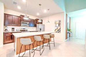A modern kitchen with dark wood cabinets, stainless steel appliances, a central island with bar stools, pendant lighting, and abstract wall art. - 7BR Windsor Cay Home w/ Pool & Game Room (Clermont)