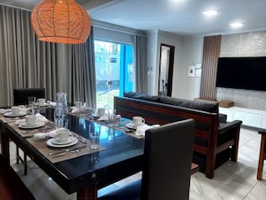 A dining area with a long table set for four, a sofa, and a television. - Cj17MSII-3D Porto Seguro Piscina/Sauna/ArConforto (Porto Seguro)