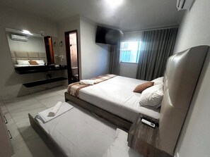 Une chambre d’hôtel comprenant un lit, une table de chevet, un miroir, un téléviseur à écran plat, une fenêtre avec des rideaux et un climatiseur fixé au mur.
