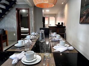 Une salle à manger avec une longue table dressée pour le repas, un escalier et un tableau accroché au mur.