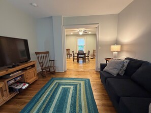 Ein Wohnzimmer mit Fernseher, einem Schaukelstuhl, einer schwarzen Couch, einem gemusterten Teppich und einem Essbereich mit Tisch und Stühlen.
