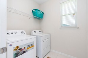 A laundry area with two white washers and dryers, a white shelf, a teal laundry basket, and a window with blinds. - Modern 7BR Villa Pool + Resort Amenities (Kissimmee)