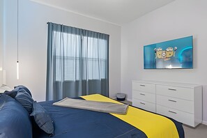A modern bedroom with a bed, a large window with sheer curtains, a white dresser, and a wall-mounted TV displaying cartoon characters. - Paradiso Grande Luxury w/ Cinema, Games & Pool (Orlando)