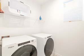 Una sala de bugaderia amb dues rentadores Whirlpool, una paret blanca, una finestra amb persianes i una estant amb un objecte blau.