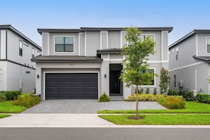 A modern two-story residential building with a garage, a small front yard with a tree, and a clear sky. - 6BR Luxury Villa Paradiso Grande Near Disney (Orlando)