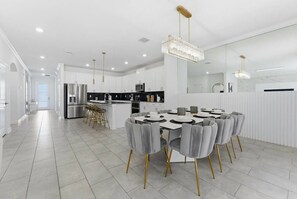 A modern kitchen with white cabinetry, a stainless steel refrigerator, and a dining area with a round table and grey upholstered chairs. - 6BR / 5BA Villa w/ Pool & Spa Storey Lake (Kissimmee)
