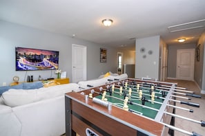 A living room with a foosball table, a sofa, a TV displaying a cityscape, and a hallway leading to another room. - 6BR Solterra Home w/ Pool + Resort Access (Davenport)