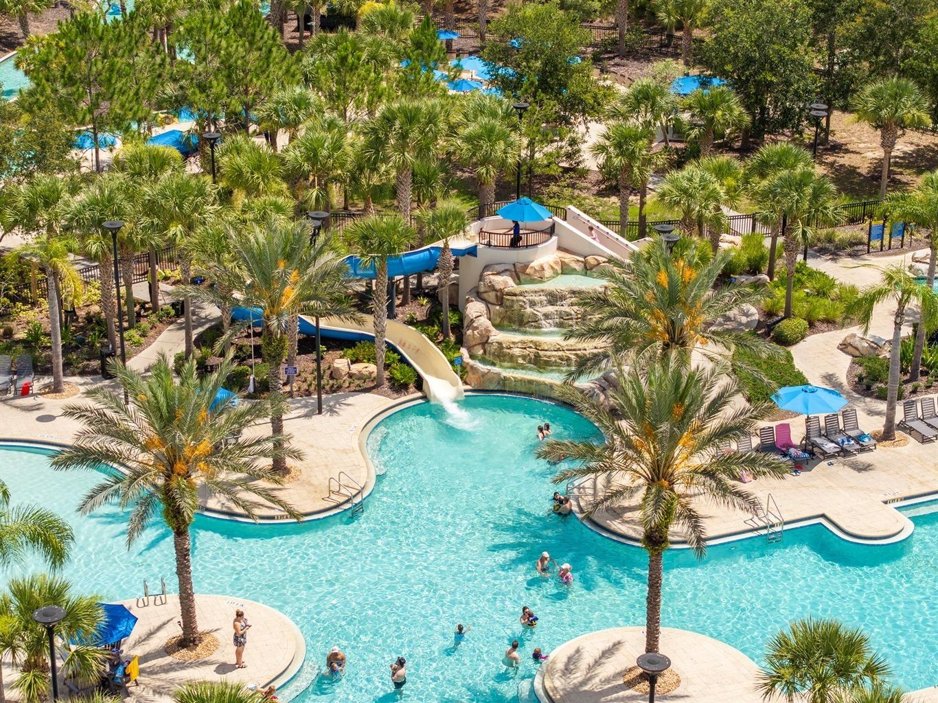 A large outdoor swimming pool with multiple water slides, surrounded by palm trees and shaded areas.