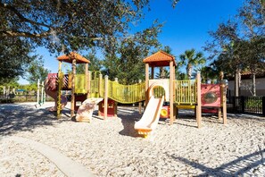 A playground with various play structures including slides and climbing areas, surrounded by trees and a clear sky. - 6BR Solterra Home w/ Pool + Resort Access (Davenport)