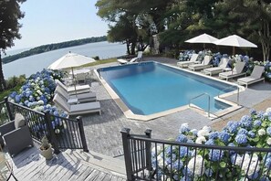 A swimming pool with lounge chairs and umbrellas, surrounded by flowers and a view of a body of water. - 4BR Waterfront house pool, hot tub, sunset view, large yard (Setauket- East Setauket)
