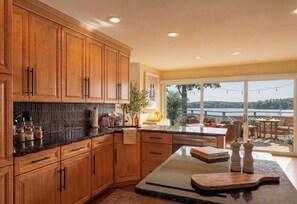 A kitchen with wooden cabinets, a granite countertop, and a view of a lake through large windows. - 4BR Waterfront house pool, hot tub, sunset view, large yard (Setauket- East Setauket)