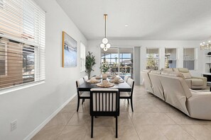 A dining area with a black table, beige chairs, and a chandelier. There is a sofa and a window with blinds in the background. - Solterra 6BR Lakeview + Pool & Game Room (Davenport)