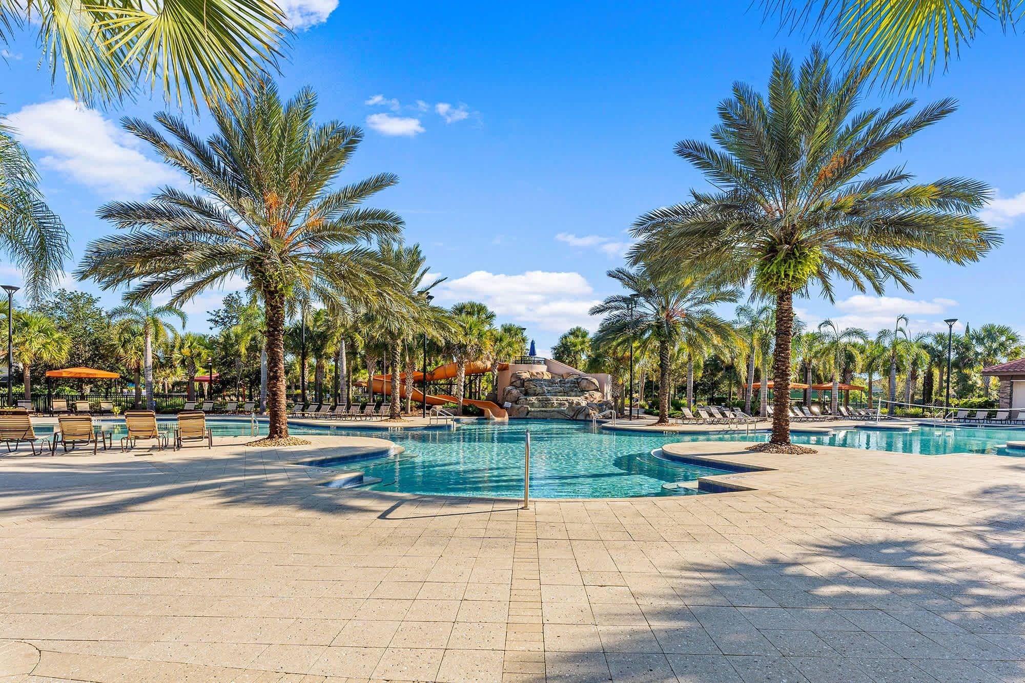 A pool area with multiple swimming pools, lounge chairs, palm trees, and a rock formation in the background.