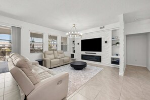A modern living room with a beige sofa, a black ottoman, and a flat-screen TV mounted on a white wall with a textured design. - Solterra 6BR Lakeview + Pool & Game Room (Davenport)