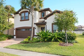 A two-story house with a brown garage door, a small porch, and a well-maintained lawn with various plants. - 8BR Home for 18 w/ Private Pool Windsor (Kissimmee)