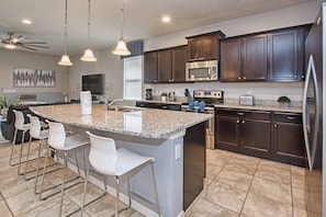 A modern kitchen with dark wood cabinets, a granite countertop, stainless steel appliances, and a bar area with white bar stools. - 8BR Home for 18 w/ Private Pool Windsor (Kissimmee)