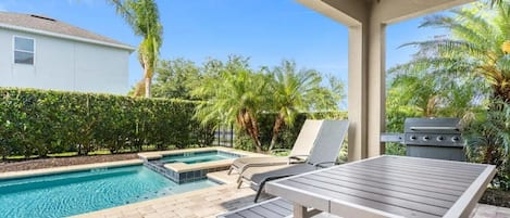 A pool area with a lounge chair, a barbecue grill, and a house in the background.