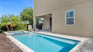 A rectangular swimming pool with a tiled deck, a covered patio area with a ceiling fan, and a view of a palm tree and a building in the background. - 7BR Themed Villa w/ Pool 6mi Disney (Kissimmee)